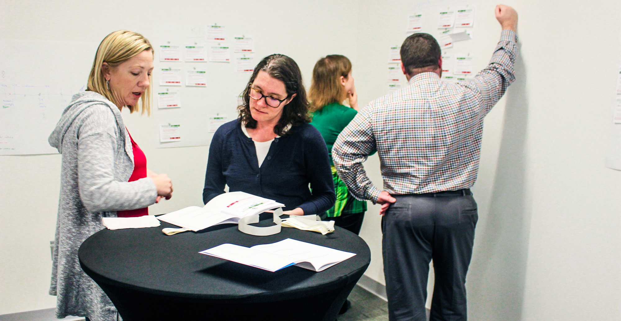 Photo of workshop attendees standing around UX strategies and sticky notes building the action plan best for their situation.